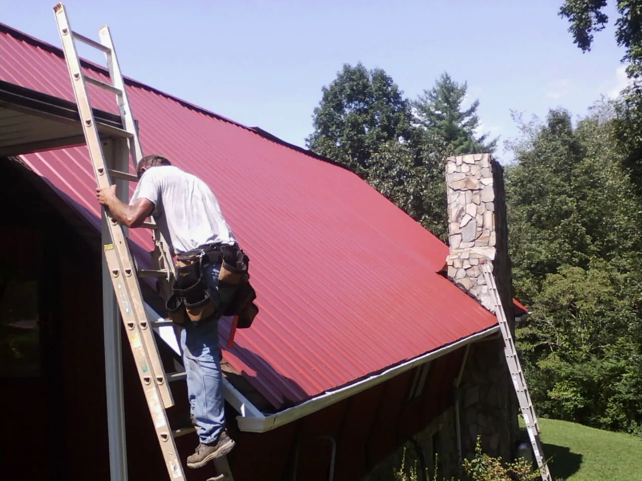 Roofer on a ladder beside a red metal roof and stone chimney — Soffit & Fascia Repair by Roofing Brothers in Laketown